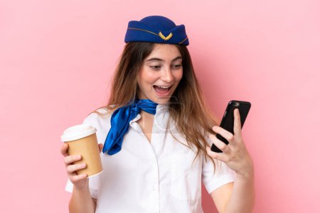 Téléchargez les photos : Airplane stewardess caucasian woman isolated on pink background holding coffee to take away and a mobile - en image libre de droit