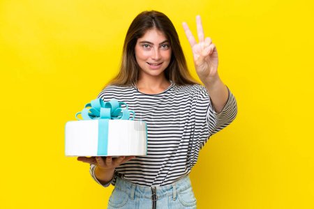 Téléchargez les photos : Jeune femme caucasienne tenant gâteau d'anniversaire isolé sur fond jaune souriant et montrant signe de victoire - en image libre de droit
