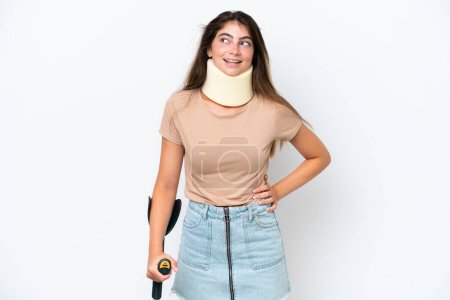 Photo for Young caucasian woman wearing neck brace and crutch isolated on white background posing with arms at hip and smiling - Royalty Free Image