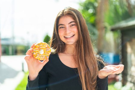 Téléchargez les photos : Jeune jolie femme caucasienne tenant un beignet à l'extérieur avec expression faciale choquée - en image libre de droit
