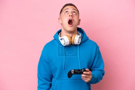 Téléchargez les photos : Jeune homme brésilien jouant avec un contrôleur de jeu vidéo isolé sur fond rose regardant vers le haut et avec une expression surprise - en image libre de droit