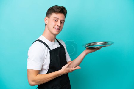 Photo for Young waitress with tray isolated on blue background extending hands to the side for inviting to come - Royalty Free Image