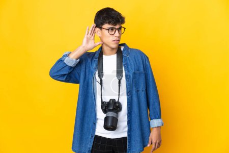 Téléchargez les photos : Jeune photographe homme sur fond jaune isolé écoutant quelque chose en mettant la main sur l'oreille - en image libre de droit
