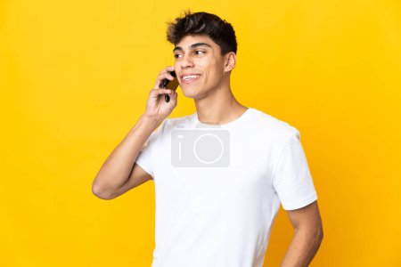 Téléchargez les photos : Jeune homme sur fond jaune isolé gardant une conversation avec le téléphone portable avec quelqu'un - en image libre de droit
