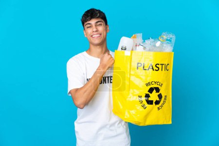 Téléchargez les photos : Tenant un sac rempli de bouteilles en plastique à recycler sur fond bleu célébrant une victoire - en image libre de droit