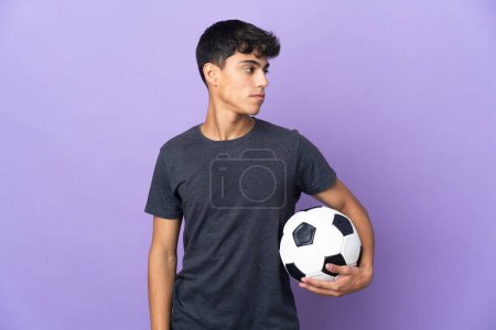 Téléchargez les photos : Jeune joueur de football homme sur fond violet isolé regardant vers le côté - en image libre de droit