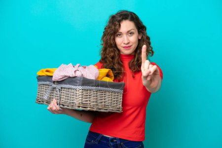 Young caucasian woman holding a clothes basket isolated on blue background showing and lifting a finger