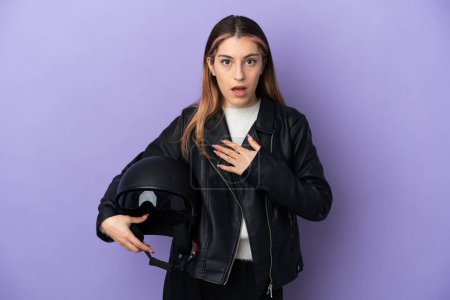 Téléchargez les photos : Jeune femme caucasienne tenant un casque de moto isolé sur fond violet surpris et choqué tout en regardant à droite - en image libre de droit