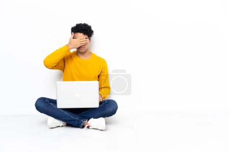 Téléchargez les photos : Homme vénézuélien assis sur le sol avec un ordinateur portable couvrant les yeux par les mains. Je ne veux pas voir quelque chose - en image libre de droit