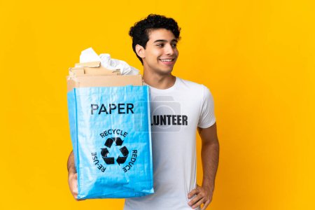 Photo for Young Venezuelan man holding a recycling bag full of paper to recycle posing with arms at hip and smiling - Royalty Free Image
