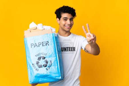 Téléchargez les photos : Jeune Vénézuélien tenant un sac de recyclage rempli de papier pour recycler souriant et montrant signe de victoire - en image libre de droit