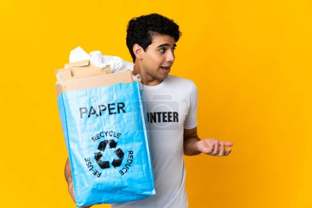 Téléchargez les photos : Jeune homme vénézuélien tenant un sac de recyclage plein de papier à recycler avec expression surprise tout en regardant côté - en image libre de droit