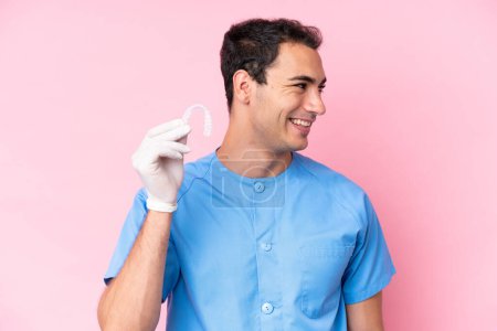 Téléchargez les photos : Dentiste caucasien homme tenant bretelles invisibles isolé sur fond rose regardant côté - en image libre de droit