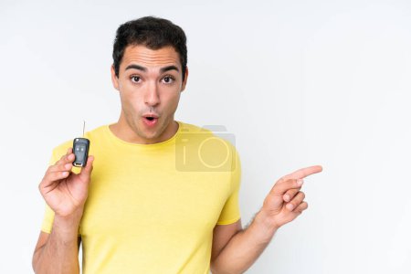 Téléchargez les photos : Jeune homme caucasien tenant des clés de voiture isolé sur fond blanc surpris et pointant côté - en image libre de droit