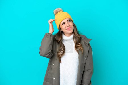 Téléchargez les photos : Jeune femme italienne portant une veste d'hiver et un chapeau isolé sur fond bleu ayant des doutes et avec une expression du visage confuse - en image libre de droit