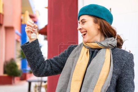 Téléchargez les photos : Femme brune tenant les clés de la maison à l'extérieur avec une expression heureuse - en image libre de droit