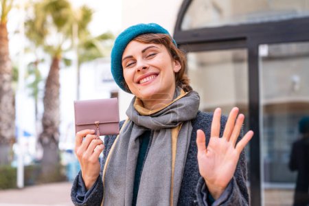 Foto de Mujer morena sosteniendo una billetera al aire libre saludando con la mano con expresión feliz - Imagen libre de derechos