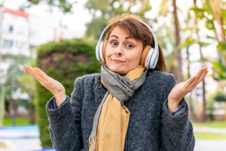 Photo for Brunette woman listening music with headphones at outdoors having doubts while raising hands - Royalty Free Image