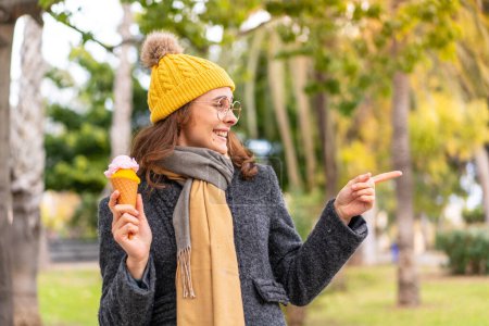 Téléchargez les photos : Brunette femme avec une crème glacée cornet à l'extérieur pointant vers le côté pour présenter un produit - en image libre de droit