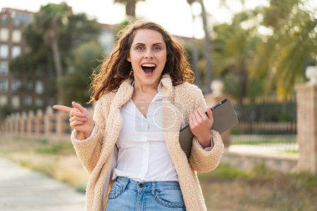Foto de Mujer joven caucásica sosteniendo una tableta al aire libre sorprendida y señalando con el dedo hacia un lado - Imagen libre de derechos