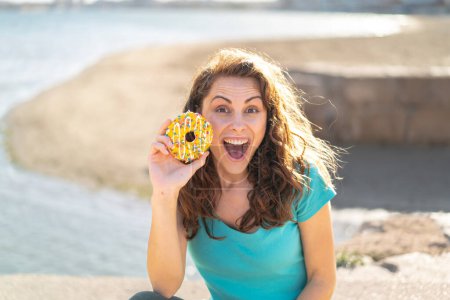 Téléchargez les photos : Jeune femme sportive tenant un beignet à l'extérieur avec surprise et expression faciale choquée - en image libre de droit