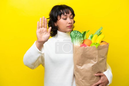 Foto de Joven argentina sosteniendo una bolsa de compras aislada sobre fondo amarillo haciendo stop gesture y decepcionada - Imagen libre de derechos