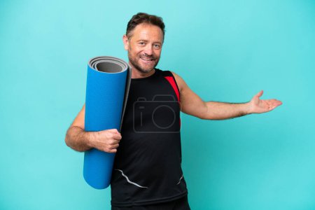 Foto de Hombre deportivo de mediana edad yendo a clases de yoga mientras sostiene una esterilla aislada sobre fondo azul extendiendo las manos hacia un lado para invitar a venir - Imagen libre de derechos