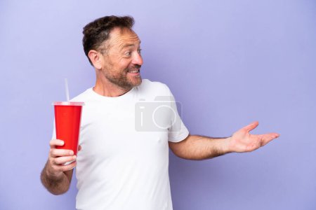 Foto de Hombre caucásico de mediana edad sosteniendo soda aislada sobre fondo púrpura con expresión sorpresa mientras mira al lado - Imagen libre de derechos