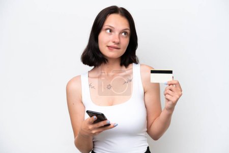 Foto de Joven mujer caucásica aislada sobre fondo blanco comprando con el móvil con una tarjeta de crédito mientras piensa - Imagen libre de derechos