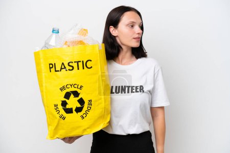 Téléchargez les photos : Jeune femme caucasienne tenant un sac rempli de bouteilles en plastique pour recycler isolé sur fond blanc regardant vers le côté - en image libre de droit