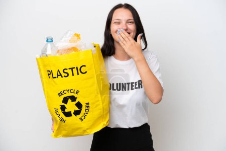 Téléchargez les photos : Jeune femme caucasienne tenant un sac rempli de bouteilles en plastique pour recycler isolé sur fond blanc heureux et souriant couvrant la bouche avec la main - en image libre de droit