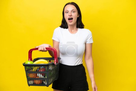 Téléchargez les photos : Jeune femme tenant un panier plein de nourriture isolée sur fond jaune avec expression faciale surprise - en image libre de droit