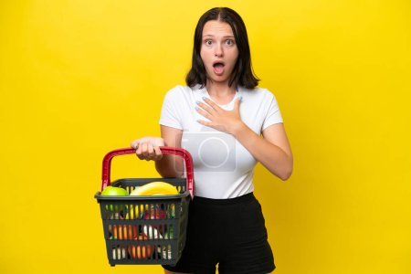 Téléchargez les photos : Jeune femme tenant un panier plein de nourriture isolée sur fond jaune surprise et choquée en regardant à droite - en image libre de droit