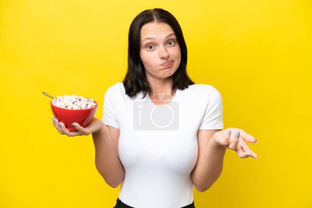Young caucasian woman holding a bowl of cereals isolated on yellow background making doubts gesture while lifting the shoulders