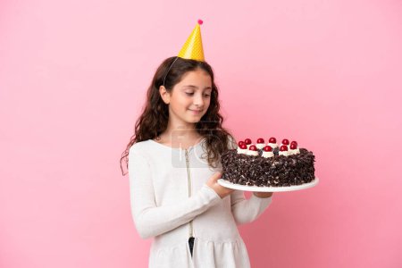 Foto de Pequeña chica caucásica sosteniendo pastel de cumpleaños aislado sobre fondo rosa con expresión feliz - Imagen libre de derechos