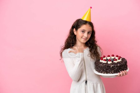 Téléchargez les photos : Petite fille caucasienne tenant gâteau d'anniversaire isolé sur fond rose pointant vers le côté pour présenter un produit - en image libre de droit