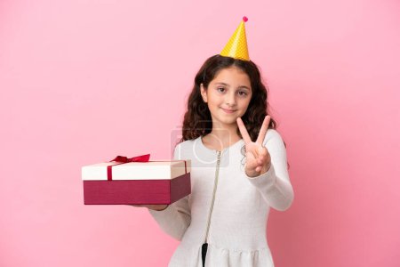 Téléchargez les photos : Petite fille caucasienne tenant un cadeau isolé sur fond rose souriant et montrant signe de victoire - en image libre de droit