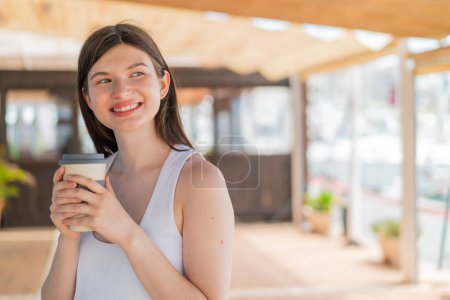 Photo for French girl with glasses holding a take away coffee at outdoors looking up while smiling - Royalty Free Image