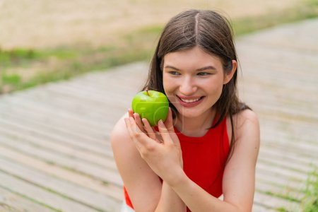 Téléchargez les photos : Jeune jolie femme à l'extérieur tenant une pomme avec une expression heureuse - en image libre de droit