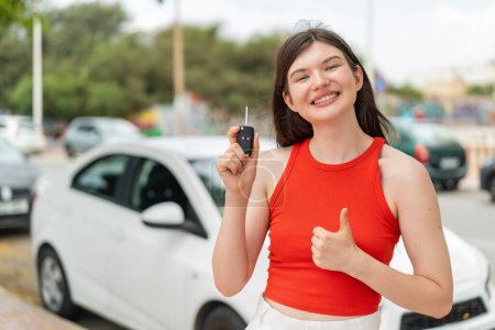 Téléchargez les photos : Jeune jolie femme ukrainienne tenant les clés de voiture à l'extérieur avec les pouces levés parce que quelque chose de bon est arrivé - en image libre de droit