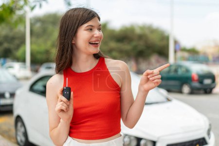 Foto de Joven bonita mujer ucraniana sosteniendo las llaves del coche al aire libre apuntando a un lado para presentar un producto - Imagen libre de derechos