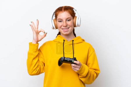 Foto de Joven mujer rojiza jugando con un controlador de videojuegos aislado sobre fondo blanco mostrando signo de ok con los dedos - Imagen libre de derechos