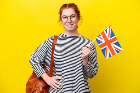 Foto de Mujer hispana joven sosteniendo una bandera del Reino Unido aislada sobre fondo amarillo posando con los brazos en la cadera y sonriendo - Imagen libre de derechos