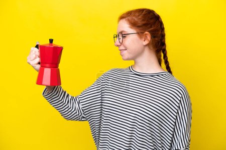 Téléchargez les photos : Jeune femme rougeâtre tenant une cafetière isolée sur fond jaune avec une expression heureuse - en image libre de droit