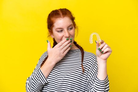 Téléchargez les photos : Jeune femme caucasienne rougeâtre tenant des bretelles invisibles isolées sur fond jaune avec une expression faciale surprise et choquée - en image libre de droit