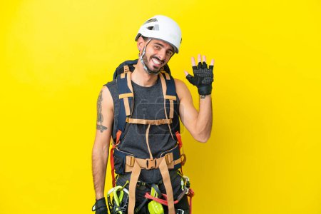 Téléchargez les photos : Jeune alpiniste caucasien isolé sur fond jaune saluant à la main avec expression heureuse - en image libre de droit
