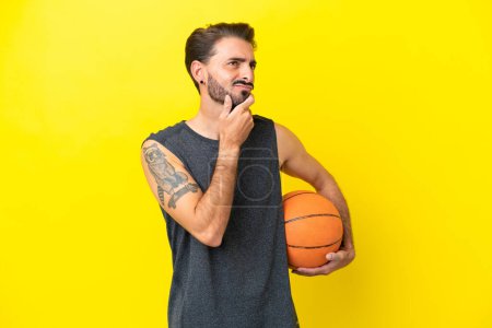 Téléchargez les photos : Beau jeune joueur de basket-ball homme isolé sur fond jaune ayant des doutes - en image libre de droit