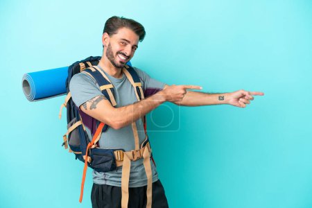 Téléchargez les photos : Jeune alpiniste homme avec un grand sac à dos isolé sur fond bleu pointant du doigt sur le côté et présentant un produit - en image libre de droit