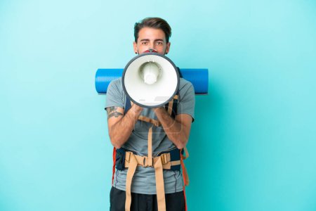 Photo for Young mountaineer man with a big backpack isolated on blue background shouting through a megaphone - Royalty Free Image