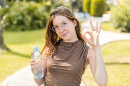 Téléchargez les photos : Française avec des lunettes tenant une bouteille d'eau à l'extérieur montrant ok signe avec les doigts - en image libre de droit